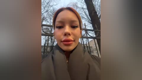 Media: A close-up video of a young Asian woman with light skin, dark hair, and a neutral expression. She wears a dark coat, standing against a background of bare trees and a metal railing.