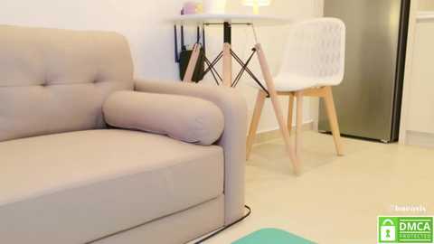 Media: Video of a minimalist living room with a beige sofa, a white chair with wooden legs, and a small round table against a white wall.