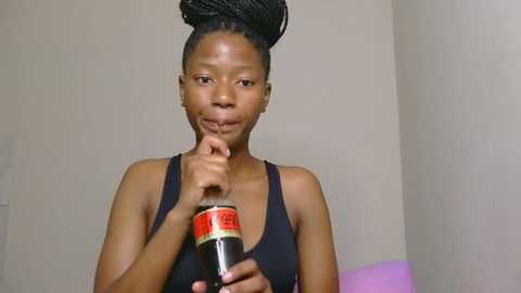 Video of a young Black woman with medium-dark skin and braided hair, wearing a black tank top, holding a bottle of Coca-Cola. She has a thoughtful expression and is touching her chin with her index finger.