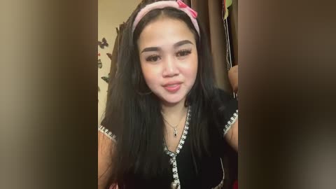 Media: Video of a young Asian woman with long black hair, wearing a pink headband, black V-neck dress with white trim, and gold necklace. Background features a brown curtain and wall decorations.