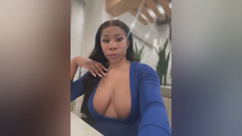 Media: Video of a Black woman with long, straight black hair, wearing a deep blue, plunging neckline dress, seated at a table in a modern, well-lit room with white walls and wooden staircase.