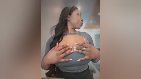 Media: Video of a black woman with long black hair, wearing a tight gray top, squeezing her large breasts with both hands. Background shows a blurred kitchen.