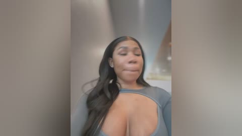 Media: A video of a Black woman with long, straight black hair, wearing a sheer gray top that reveals her large breasts, standing in an indistinct room with blurred background.