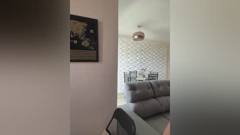 Media: Video of a modern living room with a gray sofa, a framed world map, and a dining area with black chairs and a table, featuring white geometric wallpaper and a chandelier.