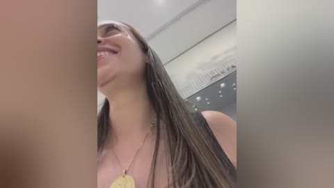 Media: A video of a smiling woman with long, wet hair, wearing a gold necklace. The background features a blurred, modern indoor setting with large windows and white walls.