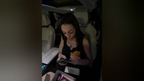 Media: A video of a woman with long black hair, wearing a black sleeveless top, seated in a dimly lit car, holding a sandwich, reading a book, with a window and interior details visible.