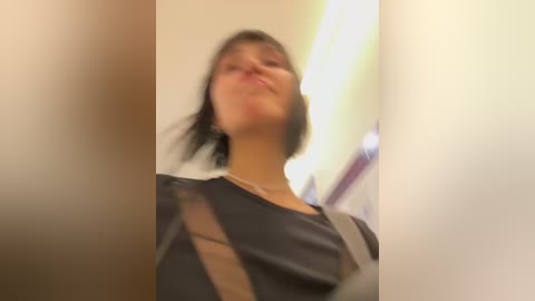 Media: A blurred video of a young woman with short dark hair, wearing a black dress with a beige shoulder strap, standing indoors against a blurred background.