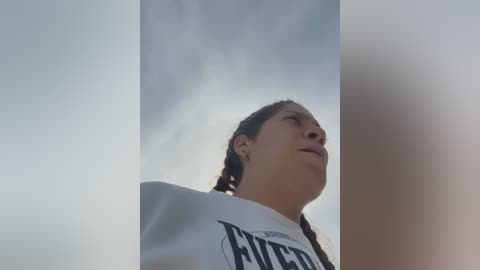 Media: A video of a young woman with a light brown complexion and braided hair, wearing a white t-shirt, looking up at the sky. The background is a cloudy sky, giving a serene, peaceful atmosphere.