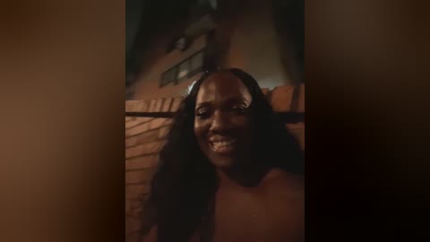 Media: Video of a joyful, dark-skinned woman with long black hair, smiling broadly, standing in a dimly lit room with brick walls and a staircase.