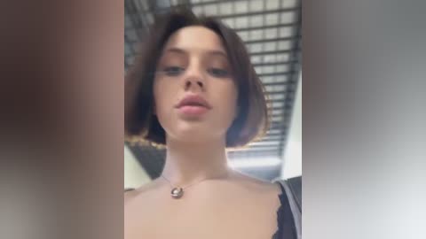 Media: Video of a young woman with light skin and short brown hair, wearing a black top with a necklace. Background features a grid ceiling and blurred objects.