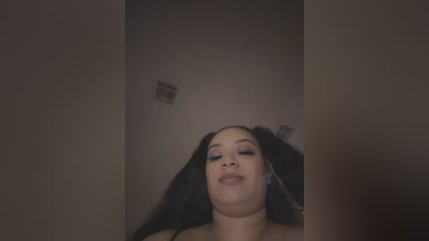 Media: Video of a smiling, light-skinned Black woman with long, straight black hair, wearing minimal makeup, in a dimly lit room with beige walls.