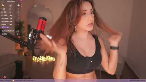 Media: Video of a light-skinned woman with long brown hair, wearing a black sports bra, standing in a dimly lit room, holding a microphone.