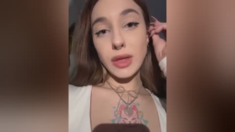 Media: Video of a young, light-skinned woman with long brown hair, wearing a white top, hoop earrings, and a colorful tattoo on her chest. Her facial features include a nose ring, full lips, and sharp eyebrows.