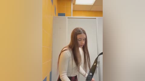 Media: Video of a young woman with long brown hair, wearing a white sweater and red plaid skirt, standing in a brightly lit, tiled bathroom with yellow and blue walls.