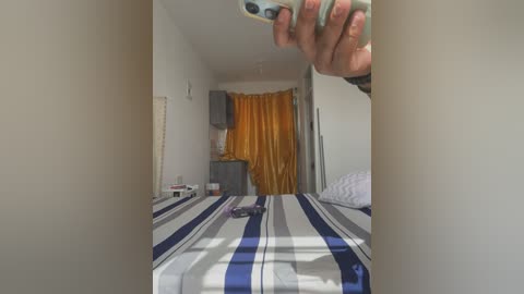 Media: Video of a simple bedroom with a bed covered in blue and white striped sheets, a mustard-yellow curtain, a white dresser, and a mirror reflecting a hand holding a smartphone.