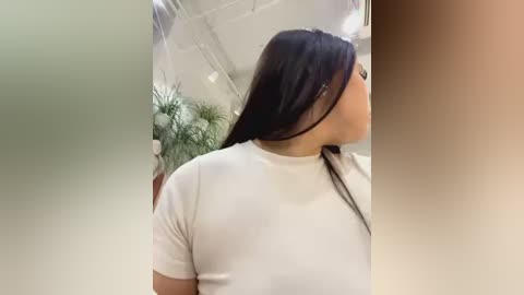 Video of a woman with long dark hair, wearing a white t-shirt, captured from the back in a modern, brightly lit indoor setting with greenery in the background.