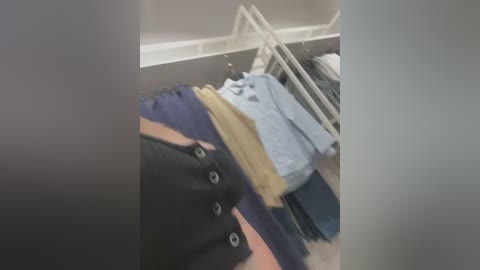 Media: Video of a person wearing a black vest over a light blue shirt, with a yellow undershirt, and blue pants, standing in a dimly lit room with a white rack of clothes in the background.