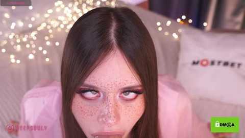 Media: Video of a young woman with fair skin, freckles, and long brown hair, wearing pink makeup and a pink jacket, in a cozy, softly lit room with twinkling fairy lights and a \"MOTEL\" pillow.