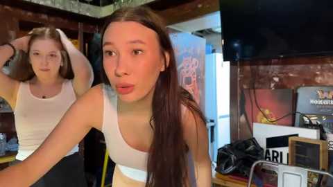 Media: A video of two young women with long hair in a dimly lit room, one adjusting her hair, wearing white tank tops, and jeans, the other in a similar outfit.