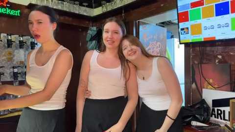 Media: Video of three young women in casual, sleeveless white tops and black skirts, laughing and posing in a lively bar with colorful posters and neon signs.