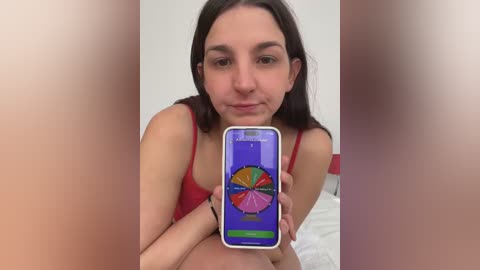 Media: A video shows a young woman with light skin and straight dark hair, wearing a red tank top, holding a smartphone displaying a colorful rainbow wheel.