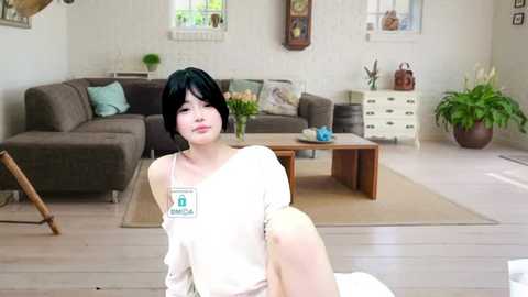 Media: Video of a young Asian woman with short black hair, wearing a white off-shoulder top, seated on a beige couch in a bright, modern living room with a brown sofa, wooden coffee table, and white brick walls.