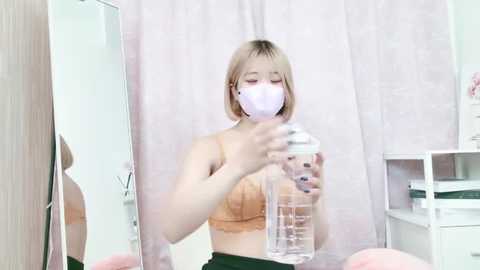 Media: Video of a young Asian woman with shoulder-length blonde hair, wearing a lavender mask, a beige bra, and black pants. She's filling a clear water bottle with a water dispenser in a bright, minimalistic room with white walls and light pink curtains.