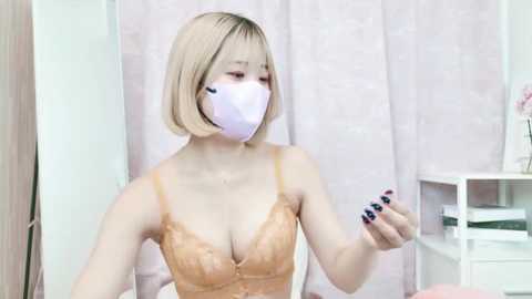 Media: Video of an Asian woman with short blonde hair, wearing a pink face mask and a beige lace bra, holding a smartphone with black nails. Background features a white shelf and pink curtains.