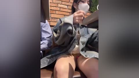 Media: Video of an Asian woman wearing a denim jacket, mask, and red panties, sitting on a wooden floor with a brick wall in the background, partially obscured by a blurred foreground.