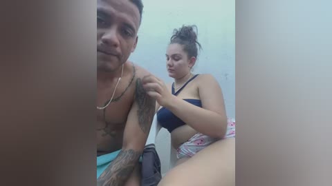 Media: Video of a shirtless, tattooed man with a gold chain, adjusting the bra of a curvy woman with dark hair in a ponytail, both in a dimly lit bathroom.