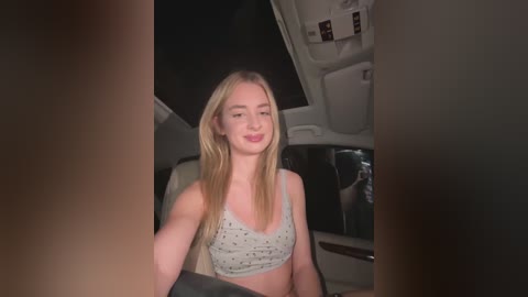 Media: Video of a blonde, fair-skinned woman with straight hair, wearing a gray sports bra, sitting in a dimly lit car at night.