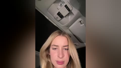 Video of a blonde woman with long hair, wearing pink lipstick, looking down at her phone, sitting on a plane seat.