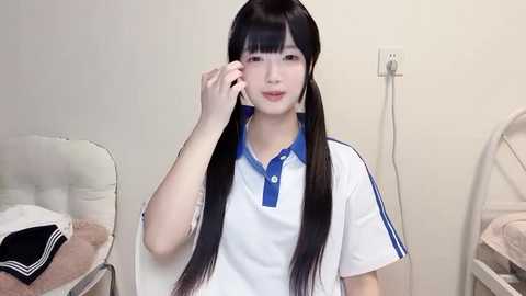 Media: Video of a young Asian woman with long black hair in pigtails, wearing a white polo shirt with blue trim, talking on a phone. She sits on a white chair in a clean, plain room with beige walls and a power outlet.