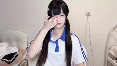 Media: Video of a young Asian woman with long black hair, wearing a white polo shirt with blue stripes and a blue collar. She has her hand over her right eye, looking distressed. Background shows a white chair and a white power outlet.