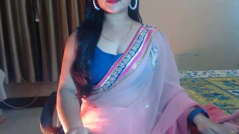 Media: Video of a South Asian woman in a pink sari with intricate gold embroidery, wearing a blue blouse, large hoop earrings, and a gold necklace, in a simple indoor setting.