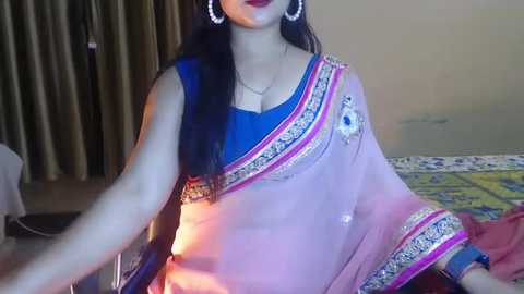 Media: A video of a woman in a blue blouse and pink sari with intricate embroidery, wearing large hoop earrings, seated on a bed with floral bedding, in a room with beige walls and curtains.