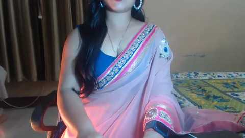 Media: Video of a South Asian woman in a pink sari with intricate blue and gold embroidery, seated indoors. She has long dark hair, wears silver earrings, and a necklace. Background shows beige curtains and a patterned bedspread.