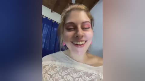 Media: A video of a young woman with light skin, blonde hair, and a white lace top, smiling with closed eyes, standing in a room with blue curtains and wooden ceiling beams.