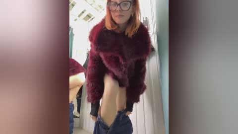 Media: Video of a slim, fair-skinned woman with shoulder-length orange hair, wearing black-framed glasses, a maroon faux fur jacket, and unbuttoned jeans. Background shows a white door and a blurred mirror reflection.