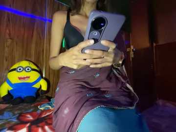 Media: Video of a young woman with fair skin, wearing a black bra and maroon skirt, holding a smartphone, standing in a dimly lit room with a Minion plush toy and a colorful blanket in the background.