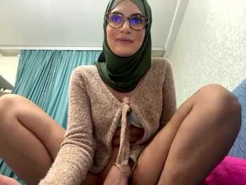 Media: A video of a young woman with a light complexion, wearing a green hijab and glasses, squatting with her legs spread, revealing her crotch. She has a beige cardigan tied at the front. The background features blue curtains and a textured wall.