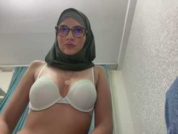 Media: Video of a young woman with light skin, wearing a green hijab, black-framed glasses, and a white lace bra, standing in a white room with teal curtains and a blue towel.