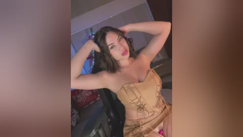 Media: Video of a young woman with fair skin and dark hair, wearing a strapless, gold corset, posing in a dimly lit room with a red and white holiday-themed pillow visible in the background.