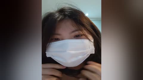 Media: Video of an East Asian woman with straight black hair, wearing a white surgical mask, partially covering her face, against a blurred indoor background.