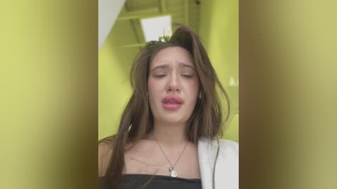 Media: Video of a young woman with long brown hair, wearing a black top, white blazer, and a silver necklace, standing indoors with yellow walls and white ceiling.