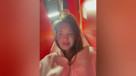 Media: Video of a young Asian woman with long brown hair and light skin, wearing a white robe, smiling in an orange-lit elevator.