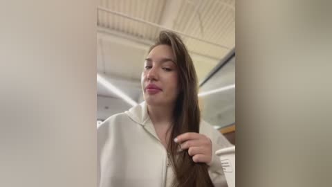 Media: Video of a young Caucasian woman with long brown hair, wearing a white blouse, smiling slightly, in a brightly lit room with a tiled ceiling and fluorescent lights.