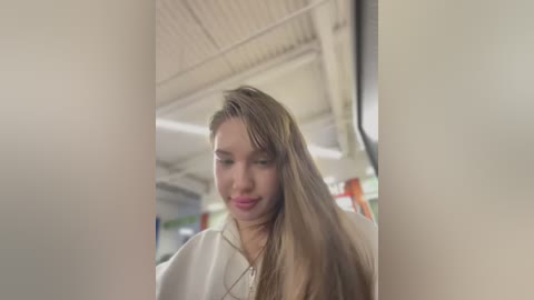 Media: A video of a young woman with long, straight brown hair and fair skin, wearing a white shirt, standing in a dimly lit, industrial-looking room with exposed pipes and fluorescent lights.