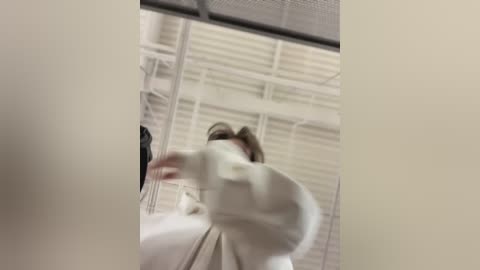 Media: A blurry video of a person wearing a white surgical mask and white gown, standing in a sterile hospital room with white walls, ceiling panels, and a window.