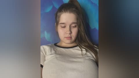 Media: Video of a young Caucasian woman with fair skin, long brown hair in a high ponytail, wearing a white ribbed t-shirt. She has a neutral expression, standing against a blue floral backdrop.